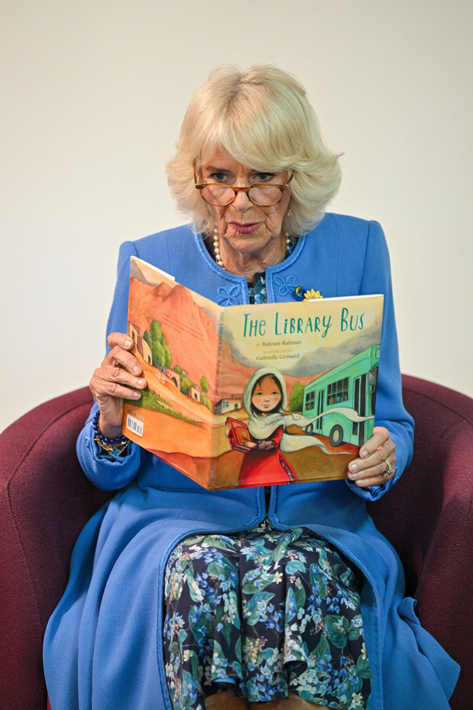 Camilla, Duchess of Cornwall, is seated in an upholstered chair while reading the picture book The Library Bus, which shows a young Afghan girl and a bus on the front cover